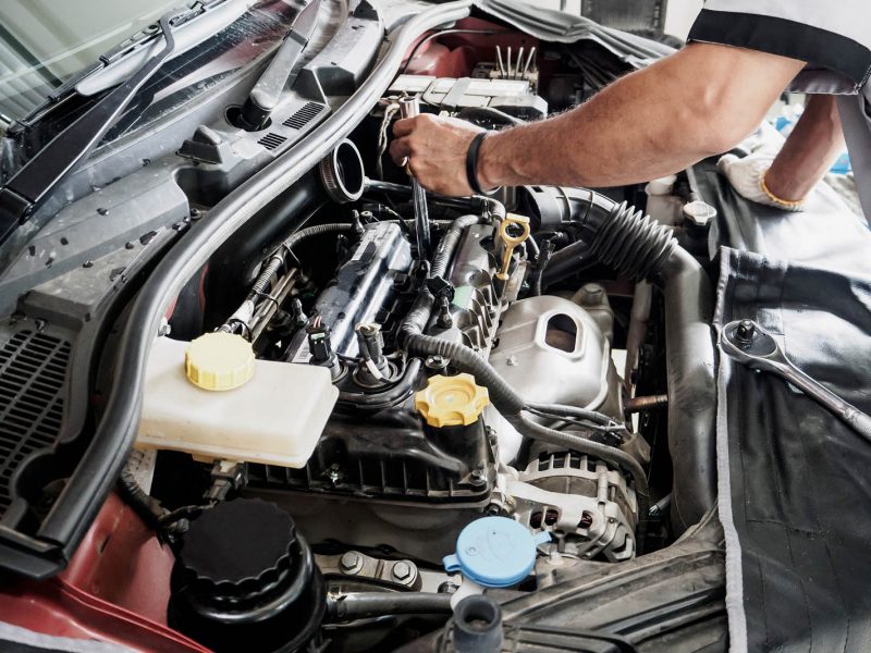 A mechanic is working on a car engine, with various tools visible. The engine parts are exposed, and the mechanic is using a wrench. The car hood is open, showing multiple components like fluid reservoirs and rubber hoses.