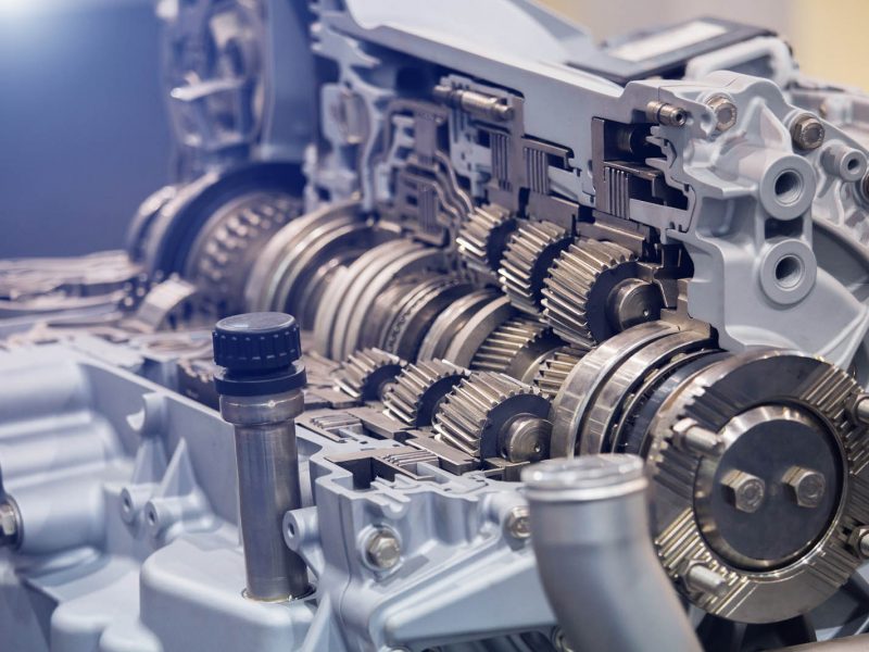 Close-up view of a mechanical assembly showcasing intricate metal gears and components, likely part of a machinery or engine system, with a mix of cylindrical and toothed structures in a silvery-grey color.