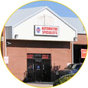 A brick building with a triangular roof displays a sign reading Automotive Specialists above the entrance. There are black glass double doors and a small logo above the sign. A car is partially visible on the right.