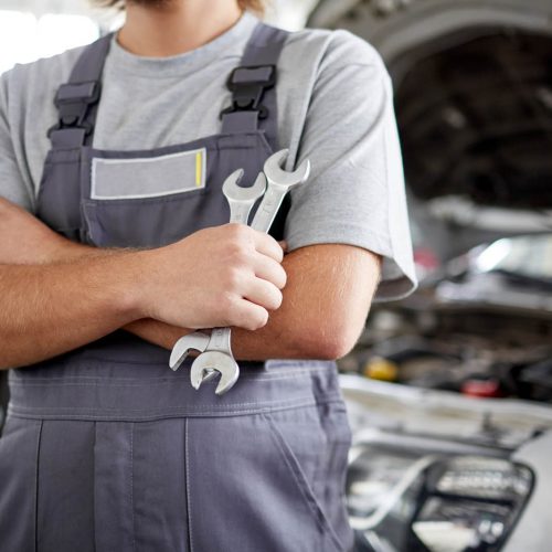 A mechanic in gray overalls stands with crossed arms holding wrenches, in front of an open car hood in a garage. Another person is blurred in the background.