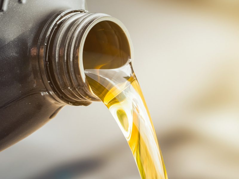 Close-up of a golden liquid being poured from a black container, likely representing motor oil. The background is blurred, highlighting the streaming liquids clarity and texture.