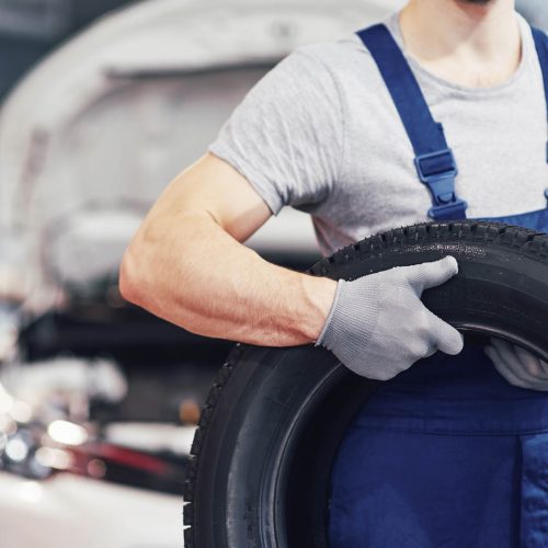 Mechanic in blue overalls and gray gloves holding a car tire in front of a vehicle with an open hood. The focus is on the tire and the mechanics upper body, with a blurred garage background.