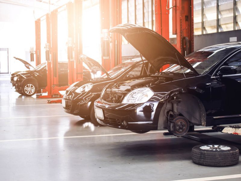 A row of cars in an auto repair shop with open hoods. The closest car is missing a wheel and is elevated on a lift. Sunlight shines through the windows, creating a bright and orderly environment.