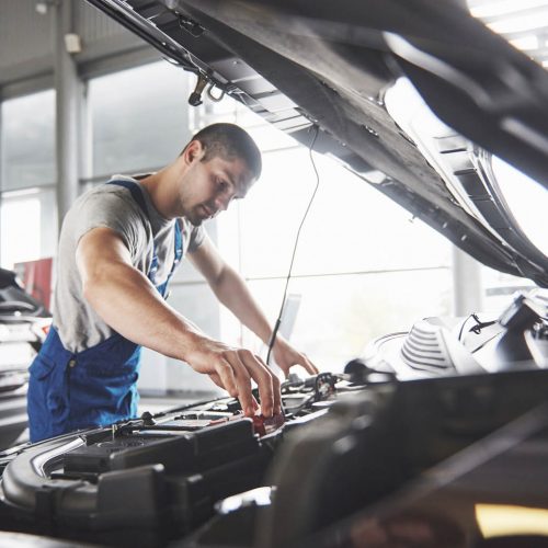 A mechanic in blue overalls and a gray shirt works under the hood of a car in a garage. The garage has large windows and another car is visible in the background.